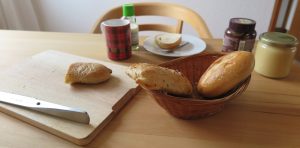 Auf einem gedeckten Frühstücks-Tisch steht ein Körbchen, in dem zwei helle Brötchen liegen. Daneben liegt ein Schneidebrett mit einem weiteren Brötchen und einem Messer. Außerdem stehen noch ein Glas Honig und Nuss-Nougat-Creme sowie eine Tasse und ein Teller daneben.